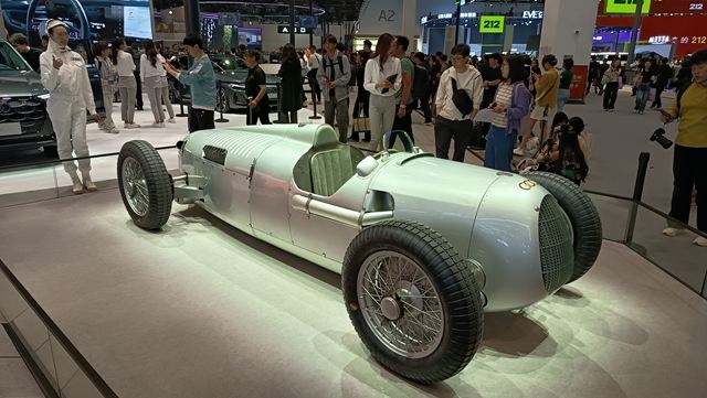 Carro esportivo antigo em exibição, com design metálico prateado e rodas grandes, cercado por uma multidão em um evento automotivo.