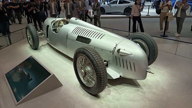 Carro de corrida vintage prateado em exibição, com pneus grandes e design aerodinâmico, cercado por espectadores em um evento de automóveis.