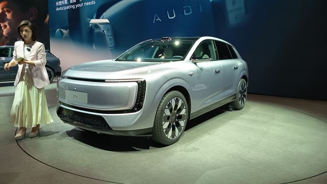 SUV moderno de cor prata em um evento automotivo, exibindo um design elegante e futurista, com uma mulher em trajes formais ao lado.
