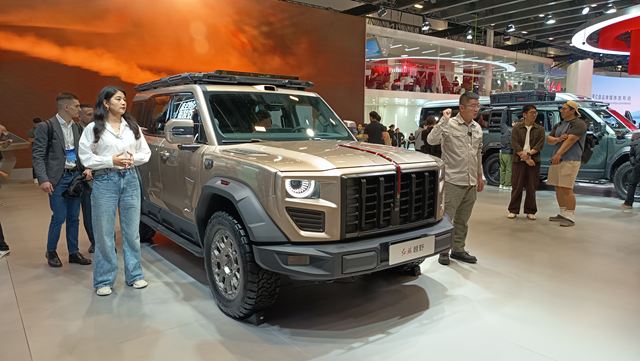 Um veículo off-road em destaque em uma feira, com pessoas ao redor observando e um apresentador mostrando o carro.