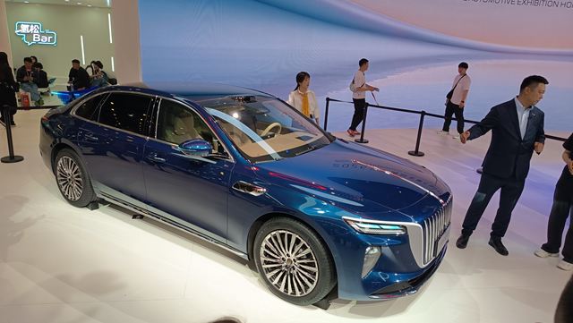 Um carro sedan azul em uma exposição automotiva, com pessoas ao fundo observando e interagindo.