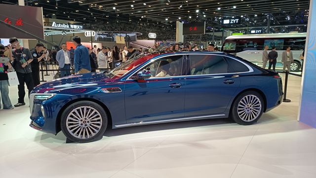 Carro sedã azul exposto em um salão do automóvel, com várias pessoas ao fundo observando.