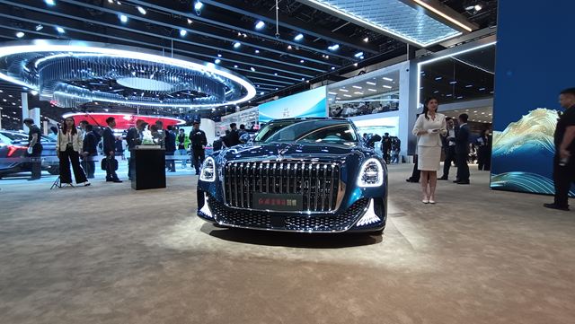 Um carro luxuoso em exibição em um salão do automóvel, com iluminação brilhante e uma multidão de visitantes ao fundo.