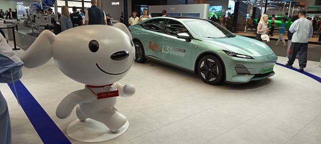 Um simpático mascote canino em formato de escultura, sorrindo, ao lado de um carro elétrico moderno em uma feira de tecnologia.