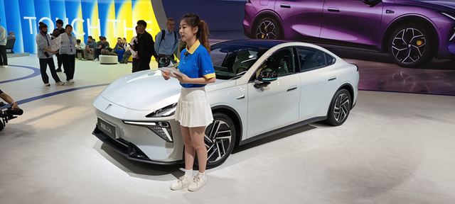 Uma apresentadora em pé ao lado de um carro elétrico branco em um evento automotivo, com um fundo colorido e público ao fundo.