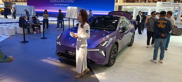 Uma mulher vestindo um traje branco interage com um carro elétrico roxo em uma feira automobilística, cercada por pessoas e exibições no fundo.
