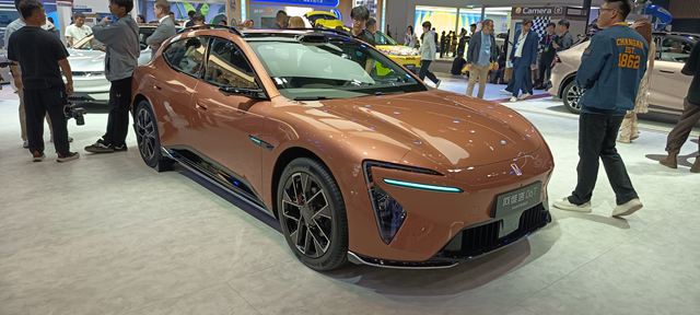 Um carro esportivo elétrico em exibição, com um design moderno e aerodinâmico, na cor laranja. O veículo está cercado por pessoas em um evento de automobilismo.
