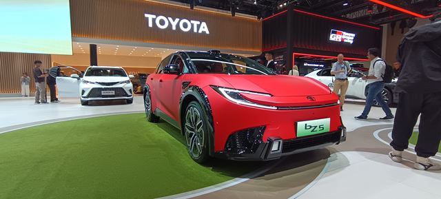 Carro elétrico vermelho da marca Toyota exposto em um salão automotivo, com um fundo de estande moderno e outras pessoas ao redor.