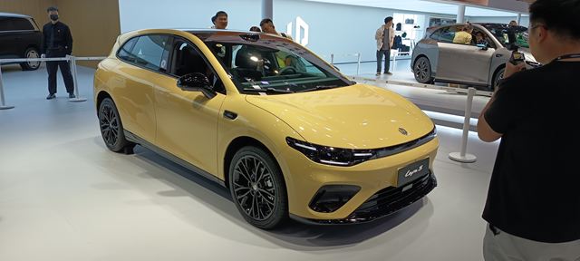 Um carro esportivo de cor amarela exibido em um salão do automóvel, com pessoas ao fundo observando e tirando fotos.