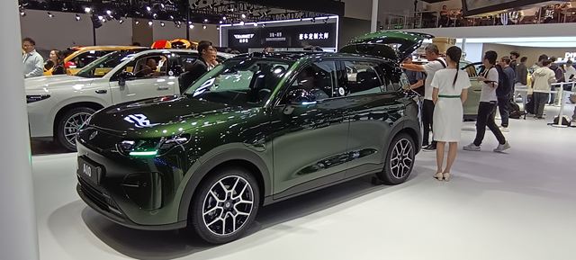 Um carro SUV verde escuro em um salão do automóvel, com pessoas interagindo ao redor e um veículo branco ao fundo.
