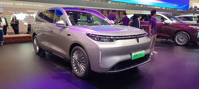 Um SUV prateado em um salão do automóvel, com pessoas ao fundo admirando o veículo.