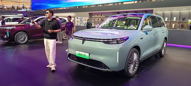 Um carro SUV elétrico em uma exposição, com um homem apresentando o veículo. O carro é de cor azul claro, com detalhes modernos e uma tela de apresentação ao fundo.