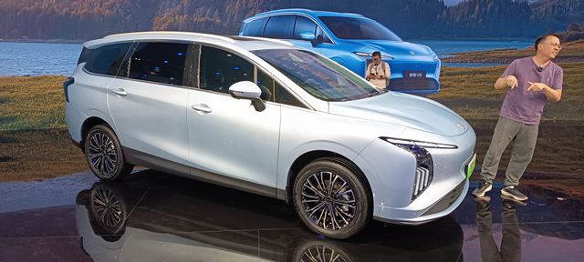 Carro da marca X apresentado em evento, com destaque para a cor prata e design moderno, ao fundo uma paisagem natural.