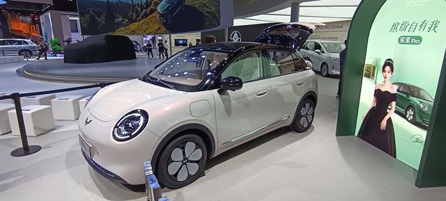 Carro elétrico compacto em exibição em um salão de automóveis, com design moderno e detalhes estilizados, incluindo rodas de acabamento em preto e branco. Ao fundo, um painel de exibição promocional com uma mulher usando um vestido elegante.