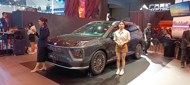 Um carro SUV cinza em um evento de exposição, com duas mulheres ao lado, uma vestindo um uniforme e a outra com uma blusa branca e saia, ambas em poses de apresentação.