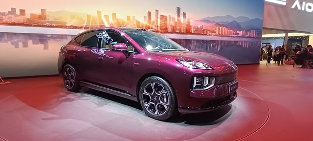 Um carro esportivo de cor roxa exposto em um salão de automóveis, com um fundo que apresenta um skyline urbano ao entardecer.