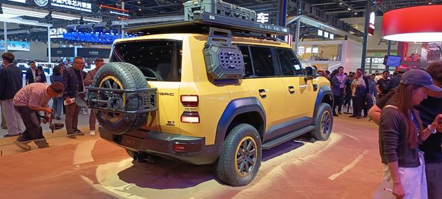 imagem de um SUV amarelo exibido em um show automobilístico, com um pneu sobressalente montado na parte traseira e detalhes em preto, cercado por visitantes.