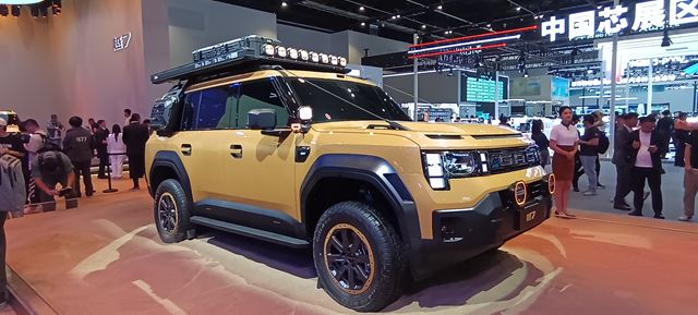 Um veículo utilitário esportivo (SUV) amarelo com design robusto, equipado com um conjunto de luzes no teto, exposto em um salão de automóveis. O fundo apresenta várias pessoas e estandes de exposições.