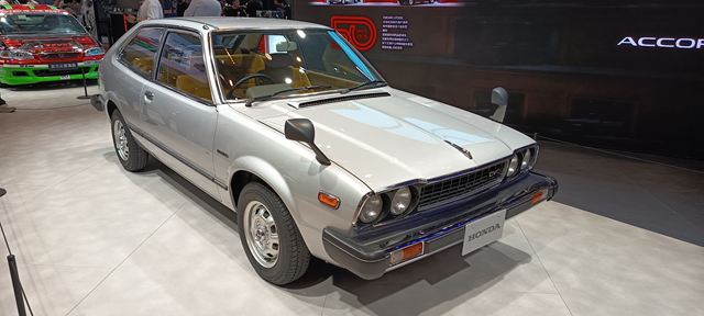Um carro compacto da Honda, modelo antigo, de cor prateada, estacionado em um evento automotivo, com detalhes do interior visíveis através das janelas.