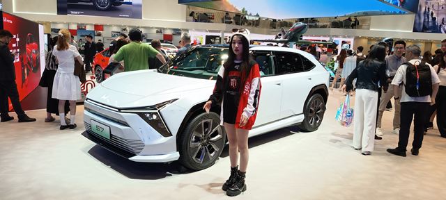 Uma jovem em pé ao lado de um carro branco em um evento, rodeada por pessoas e estandes de exposição.