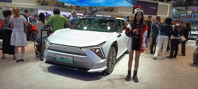 Um carro branco modelo S7 em destaque em um salão do automóvel, com várias pessoas ao fundo interagindo e observando.
