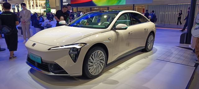 Carro elétrico de design moderno, com pintura clara e rodas distintas, exibido em um estande em ambiente urbano.