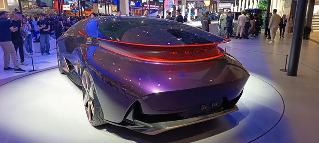 Vista traseira de um carro futurista de cor roxa, com detalhes brilhantes e luzes traseiras em destaque, em um salão automotivo lotado de pessoas.