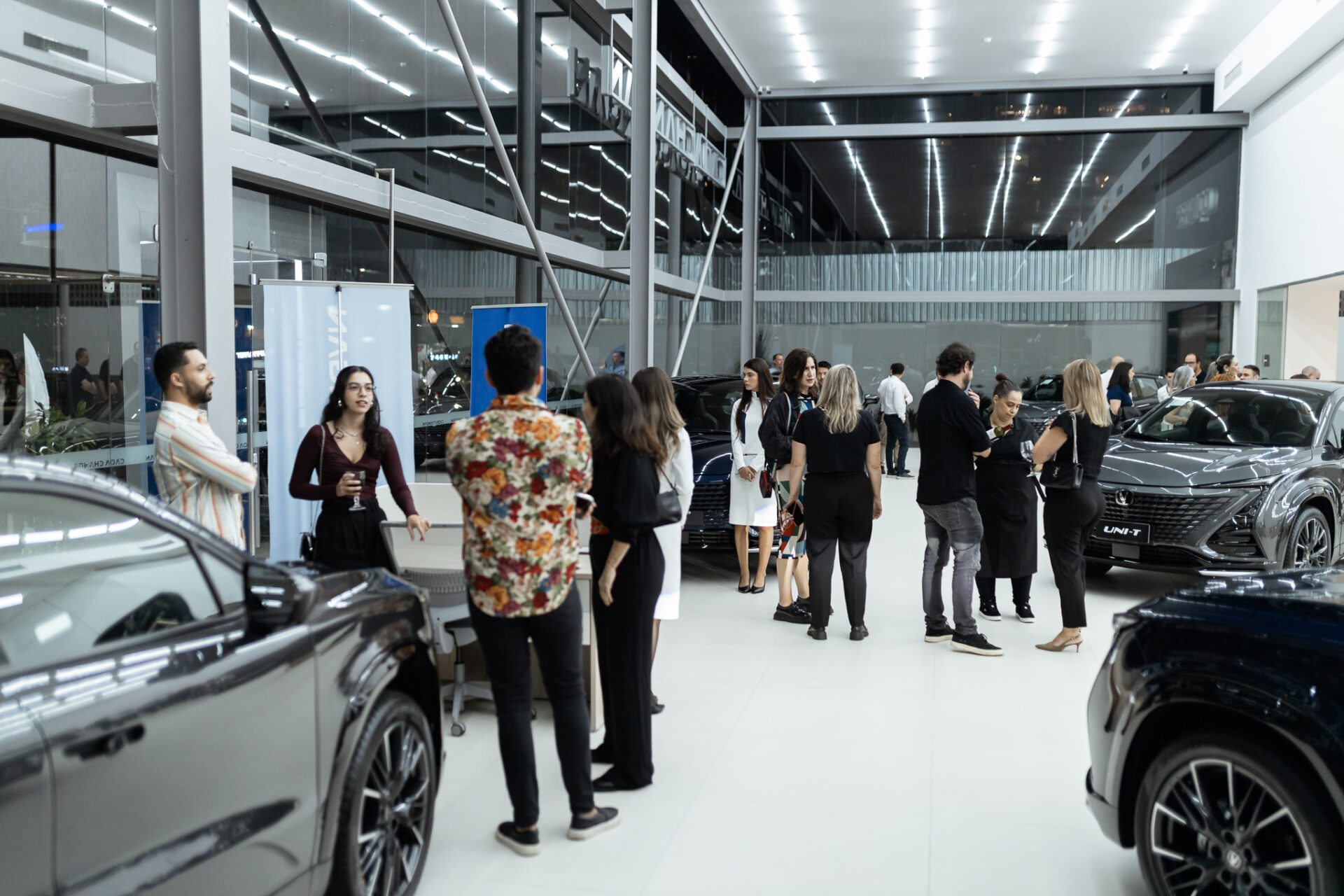 Evento de lançamento em uma concessionária de carros, com várias pessoas socializando e vários veículos expostos no interior moderno.