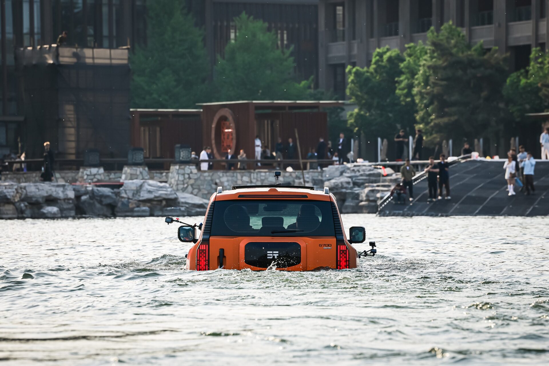 An orange SUV partially submerged in water, with people and a building in the background.