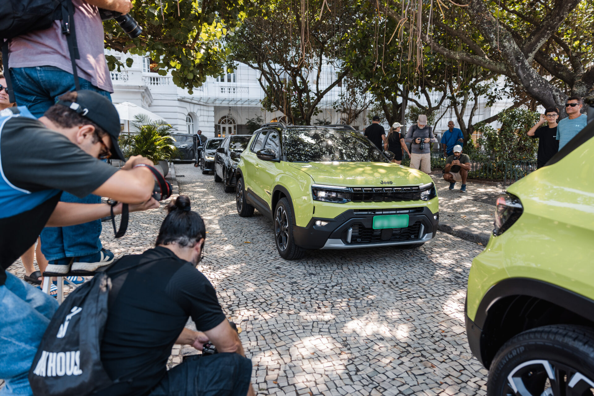 Evento de lançamento de carro com foco em dois veículos Jeep em destaque, um deles verde, cercado por jornalistas e fotógrafos em uma calçada de pedras, com árvores ao fundo.