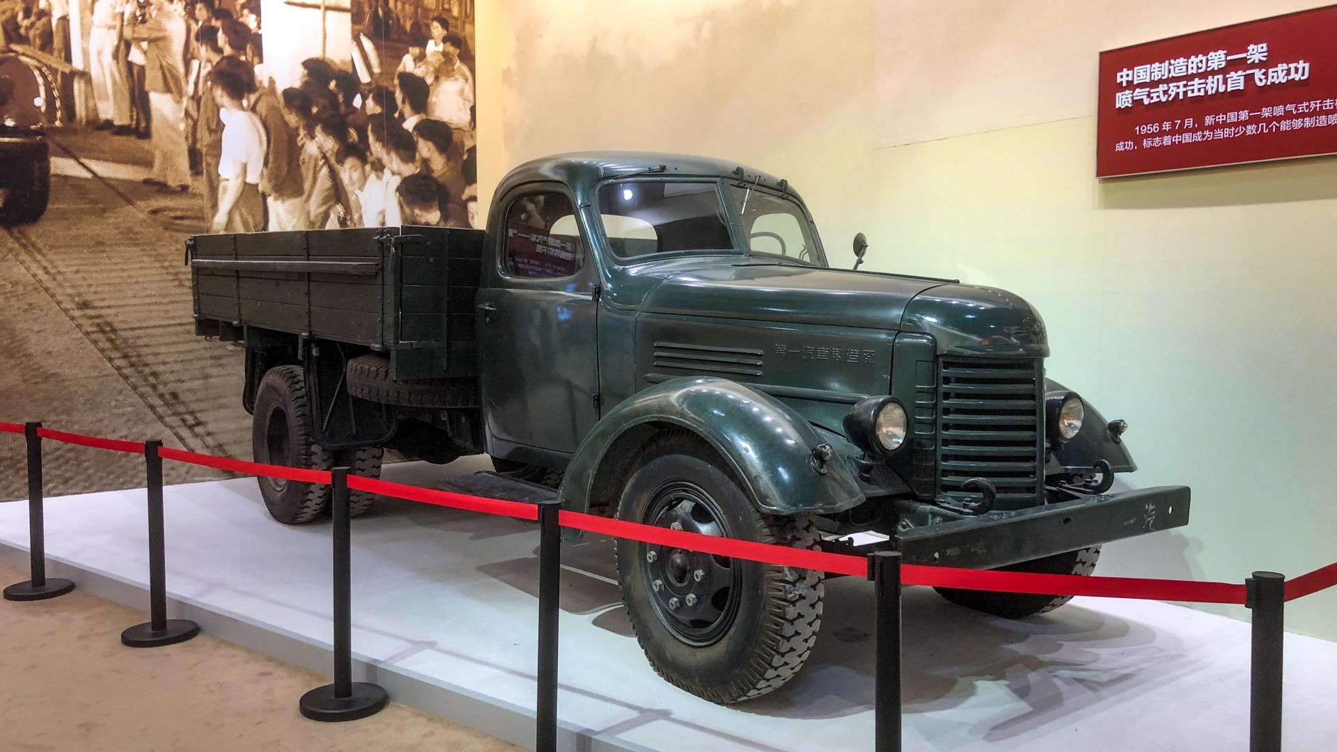 Caminhão antigo exposto em um museu, com pintura verde e carroceria de madeira, cercado por cordões de segurança.