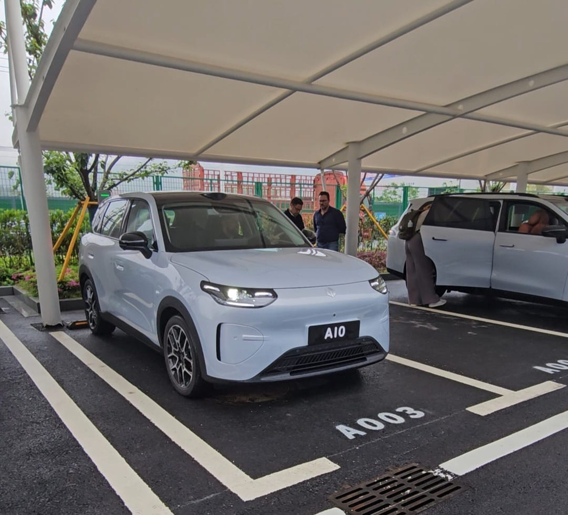 Um carro elétrico branco estacionado sob uma cobertura, com a placa A10 visível. Duas pessoas estão próximas ao veículo, enquanto outro carro branco está estacionado ao lado.