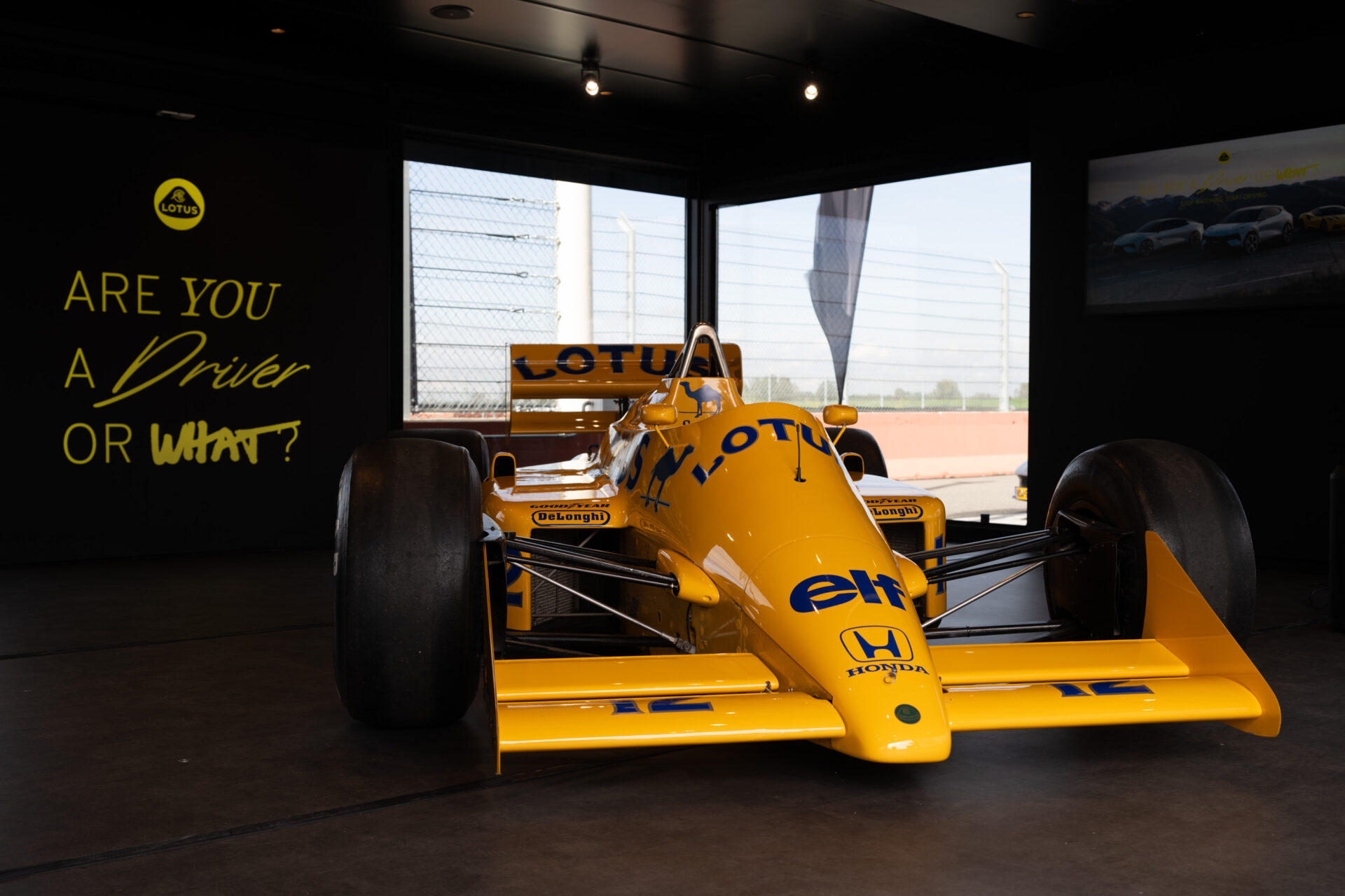 A yellow Lotus racing car displayed indoors with a minimalist background featuring the text 'ARE YOU A Driver OR WHAT?'