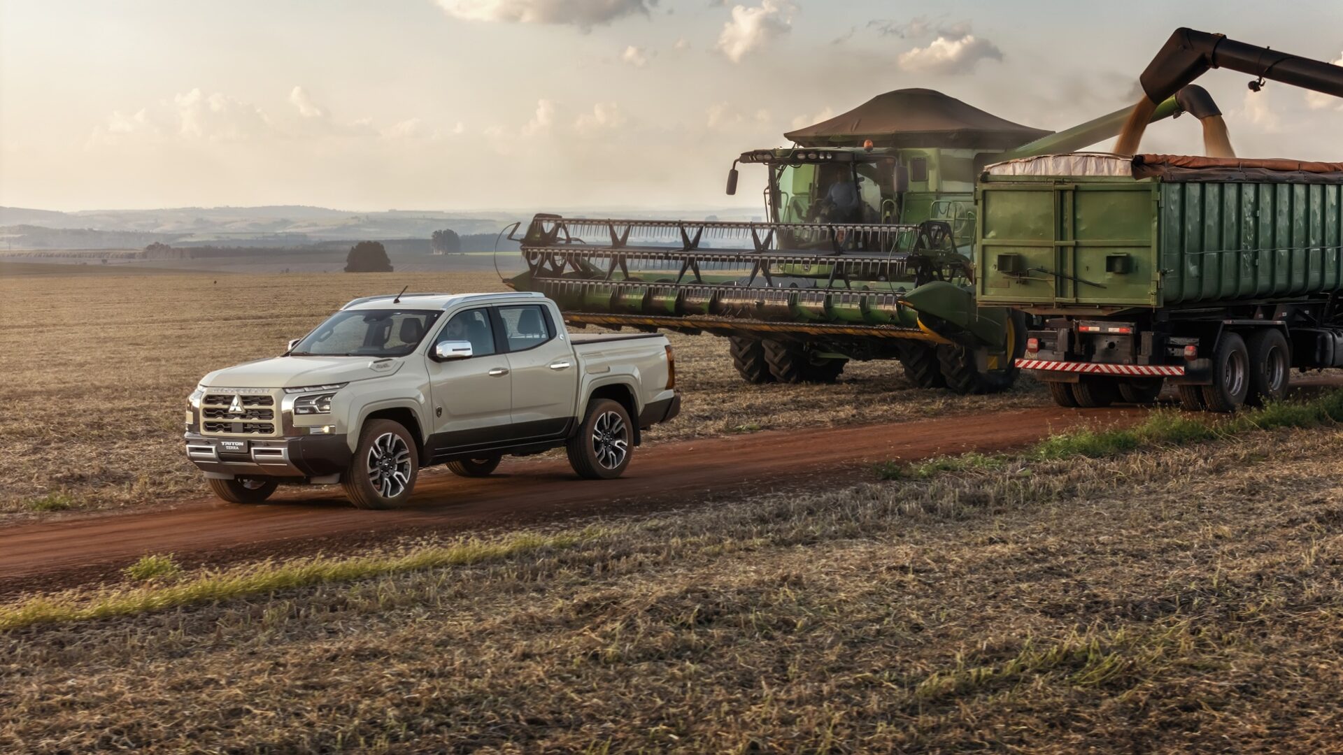 A pickup truck driving on a dirt road in an agricultural field, with a large green combine harvester and a trailer in the background under a cloudy sky.