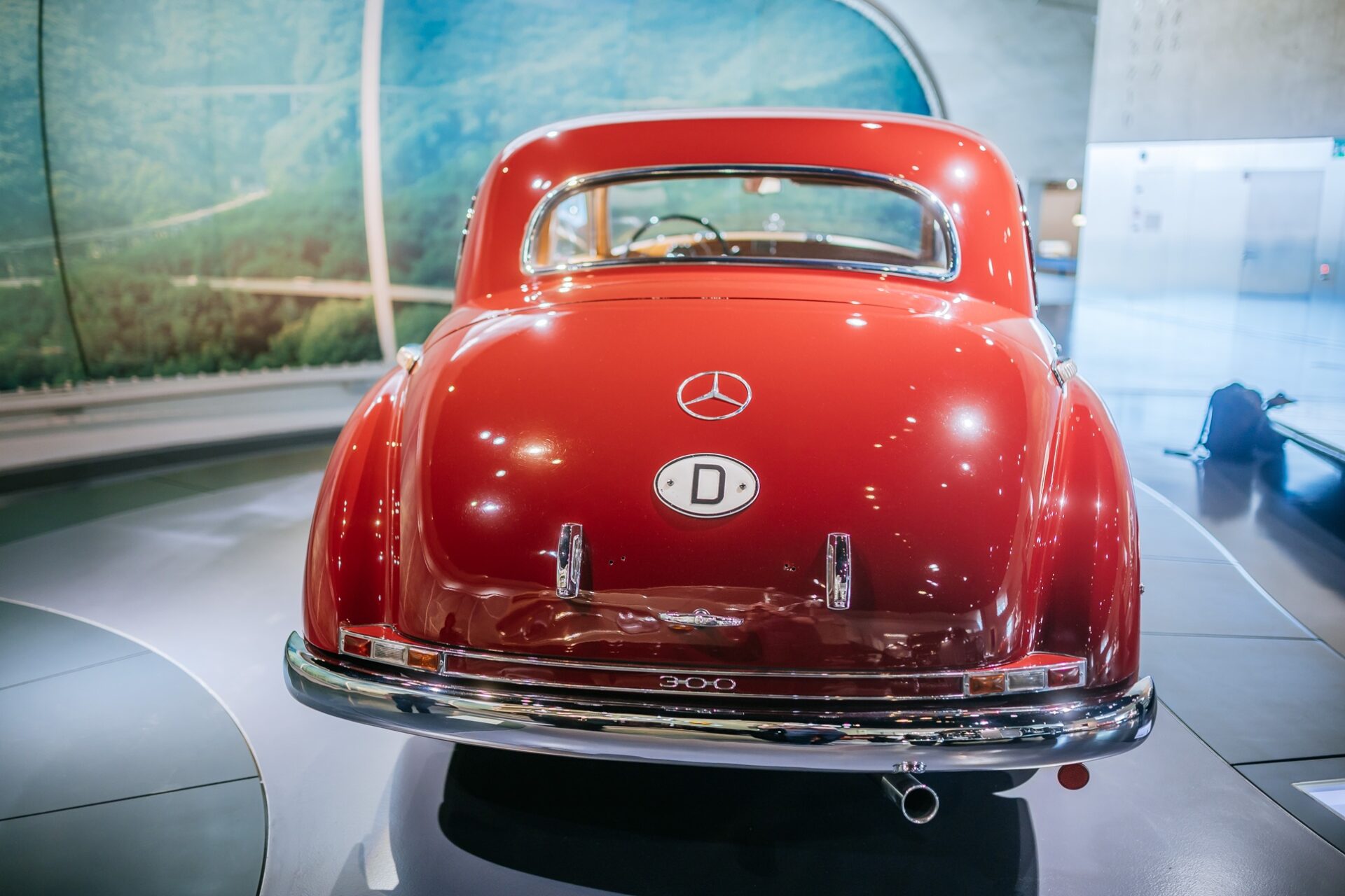 Rear view of a classic red Mercedes-Benz car, model 300, displayed in a museum setting with a scenic background.