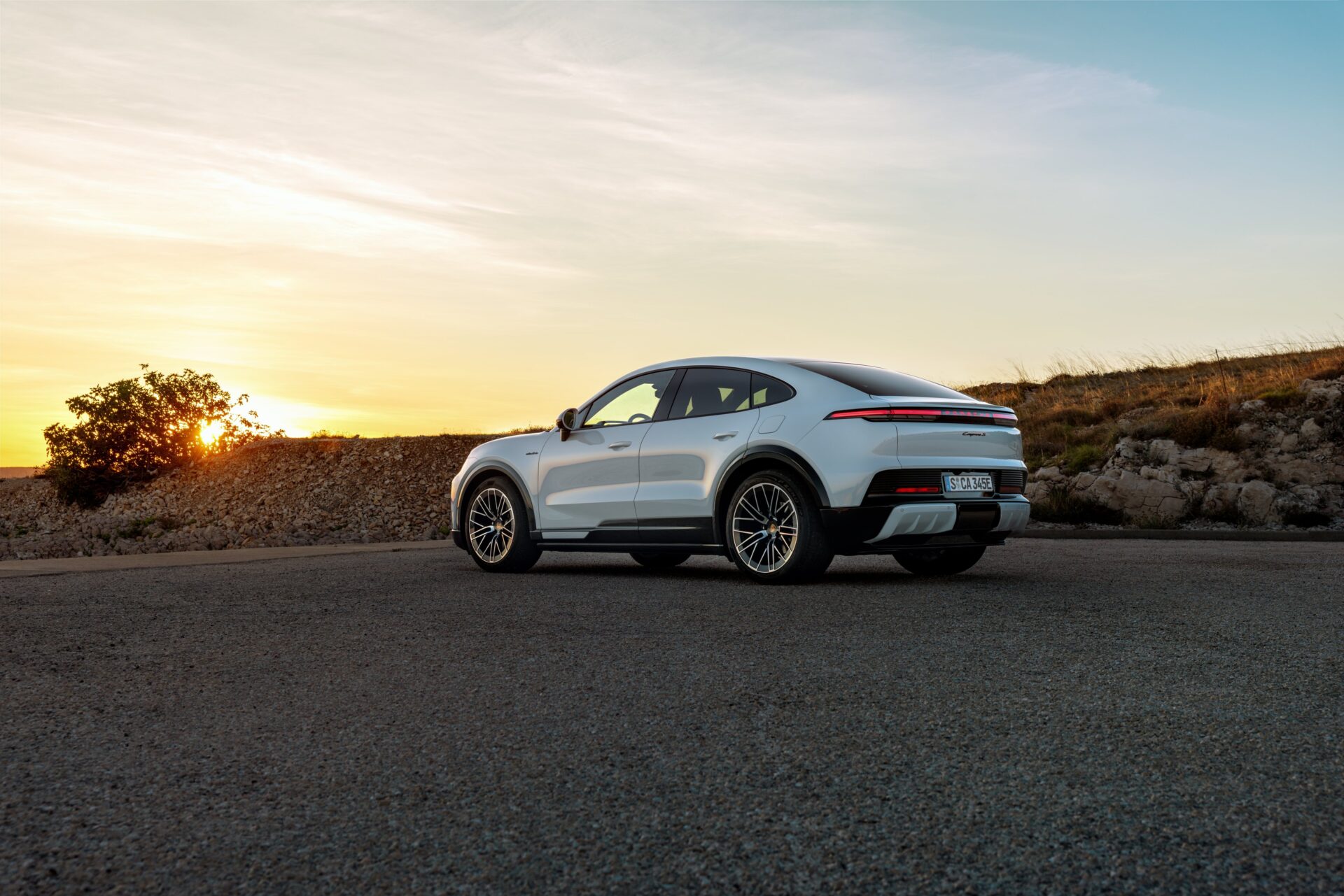 A sleek silver luxury SUV parked on a gravel road during sunset, with a clear sky and distant hills in the background.