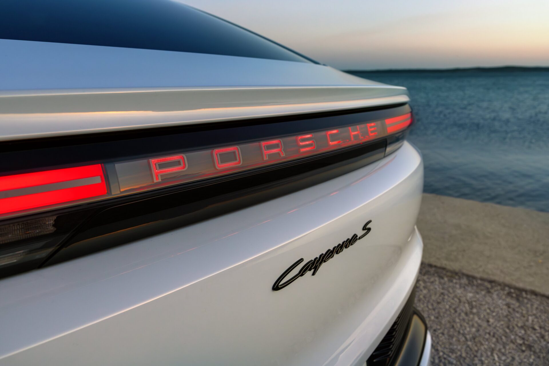 Close-up of the rear of a white Porsche Cayenne S, featuring illuminated 'Porsche' emblem and sleek design against a backdrop of water.