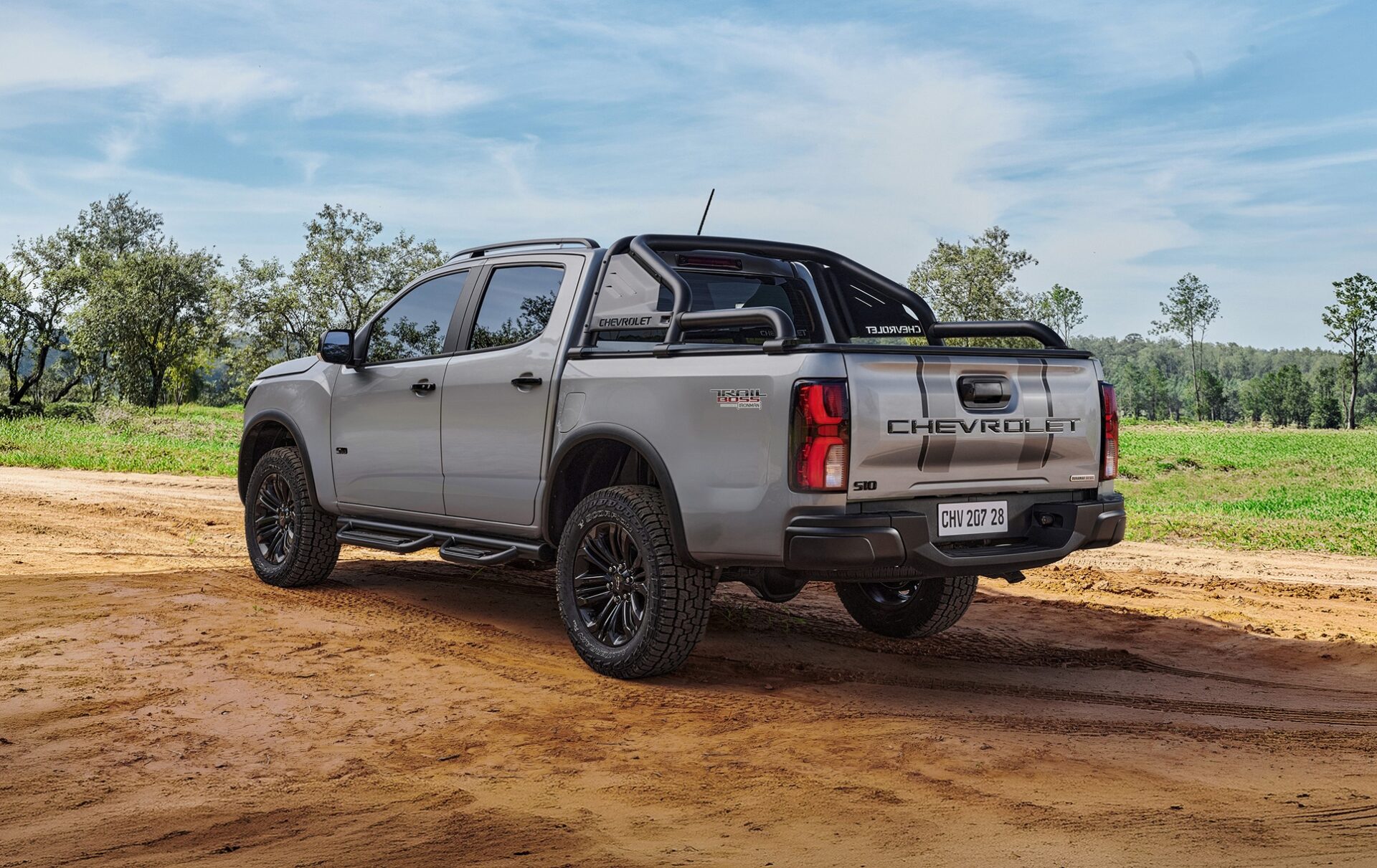 A silver Chevrolet S10 pickup truck parked on a dirt road, surrounded by greenery under a blue sky.