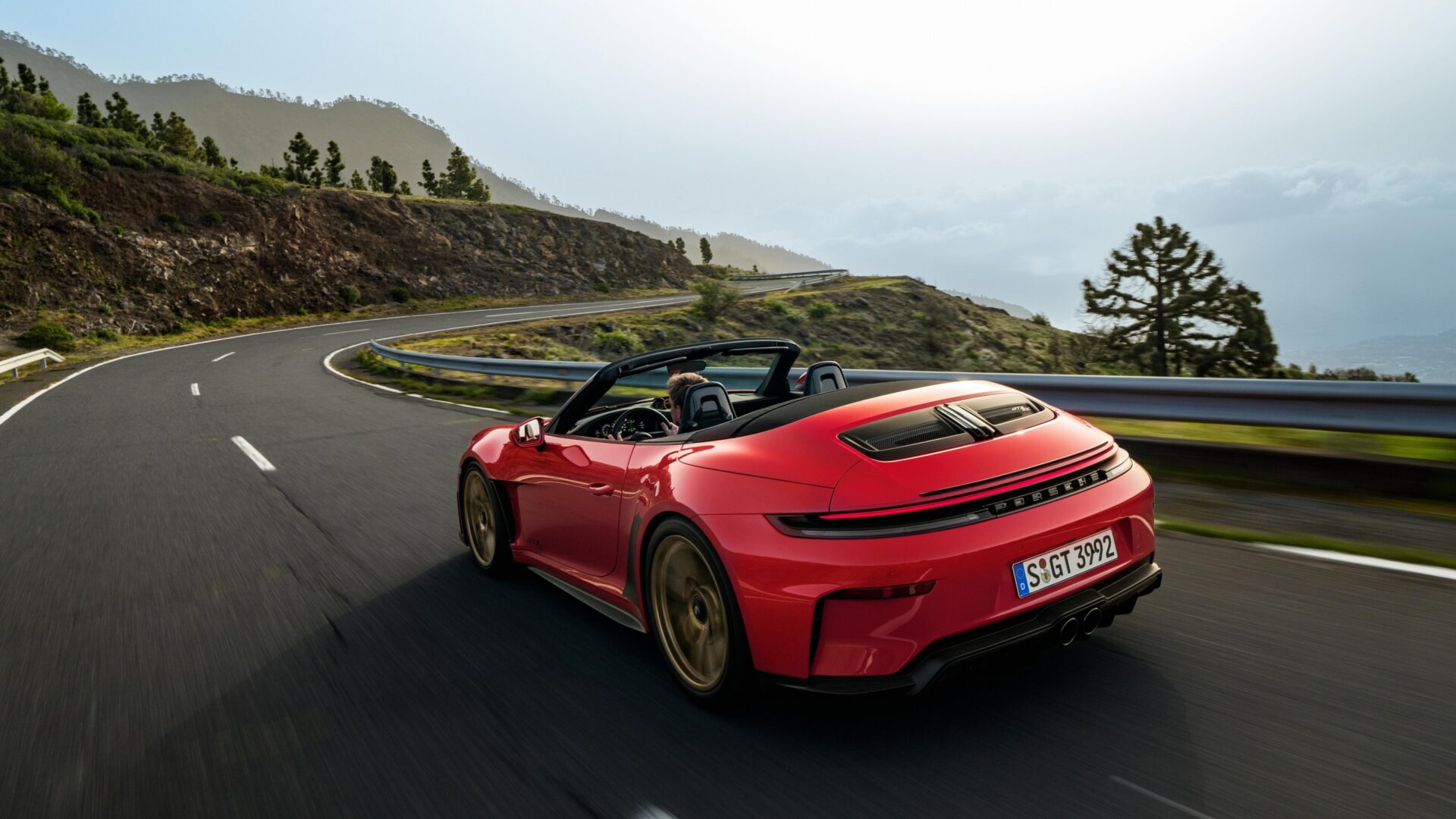 Carro conversível vermelho Porsche em uma estrada sinuosa, cercado por montanhas e vegetação.