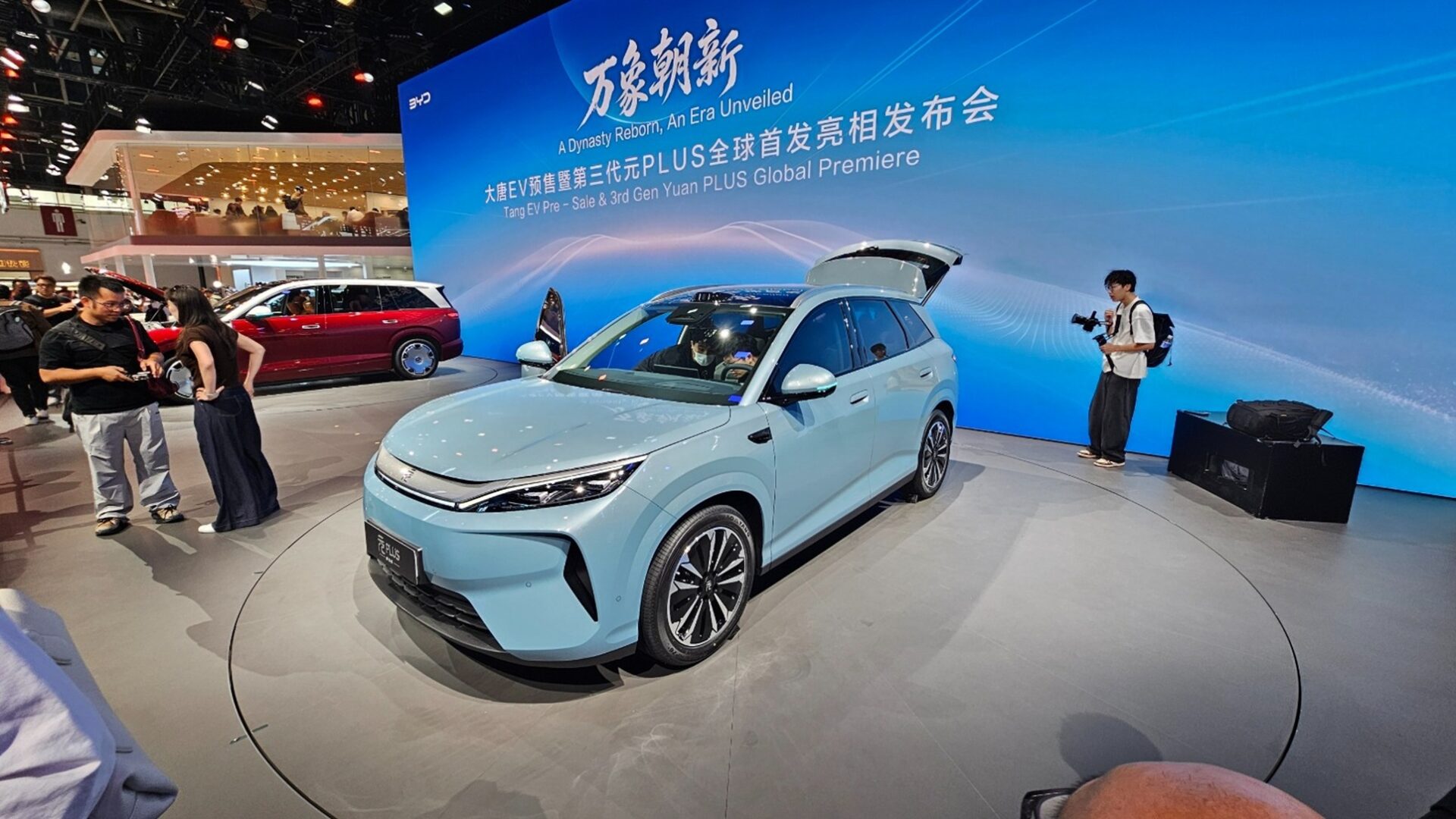 A light blue electric vehicle is displayed at a car show, surrounded by attendees. In the background, a large screen showcases the event title, and a red car is visible behind the blue vehicle.