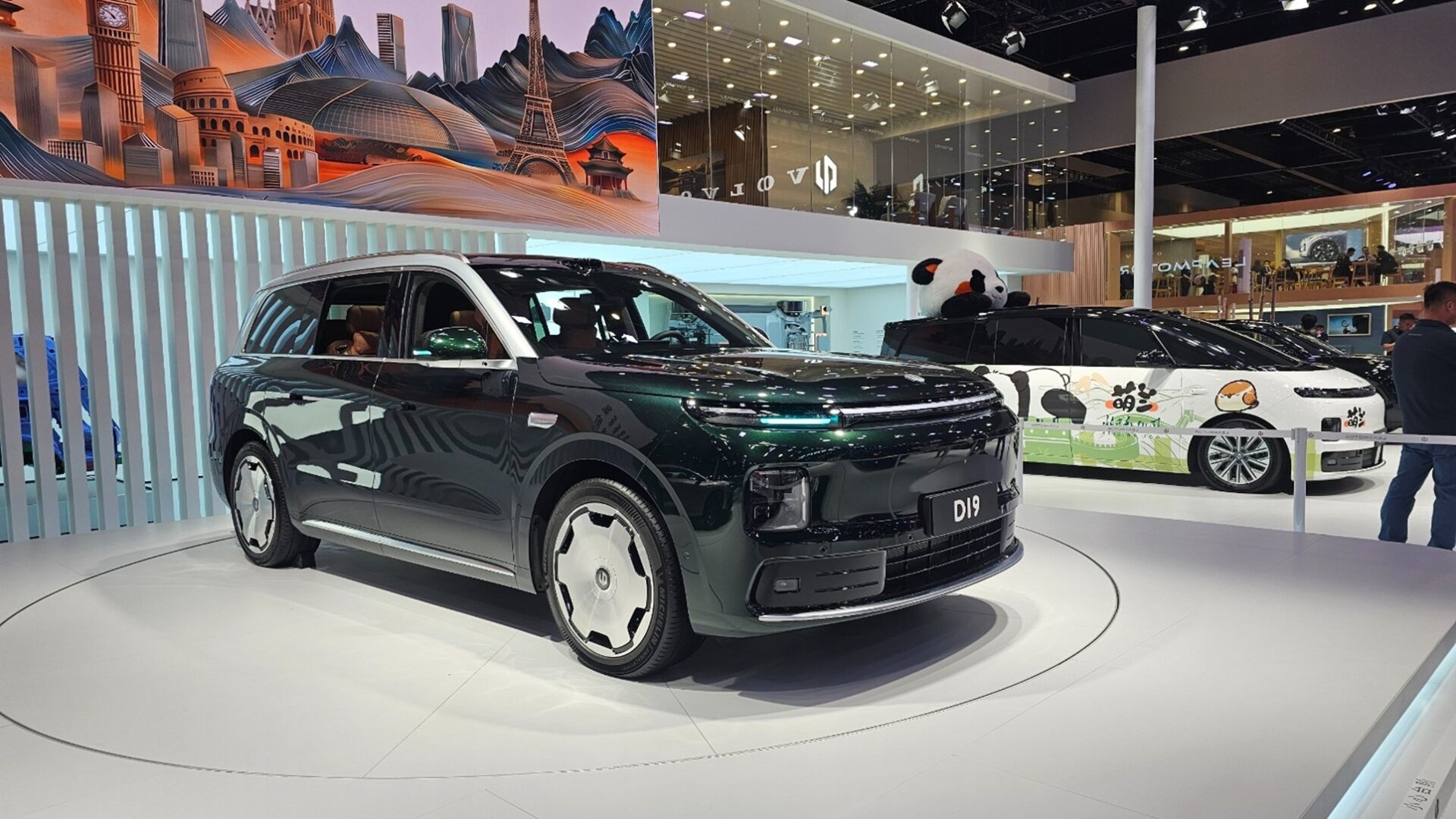 A sleek black SUV displayed at a car show, featuring modern design elements and illuminated accents. In the background, a vibrant mural depicting various landmarks and a panda-themed car are visible.