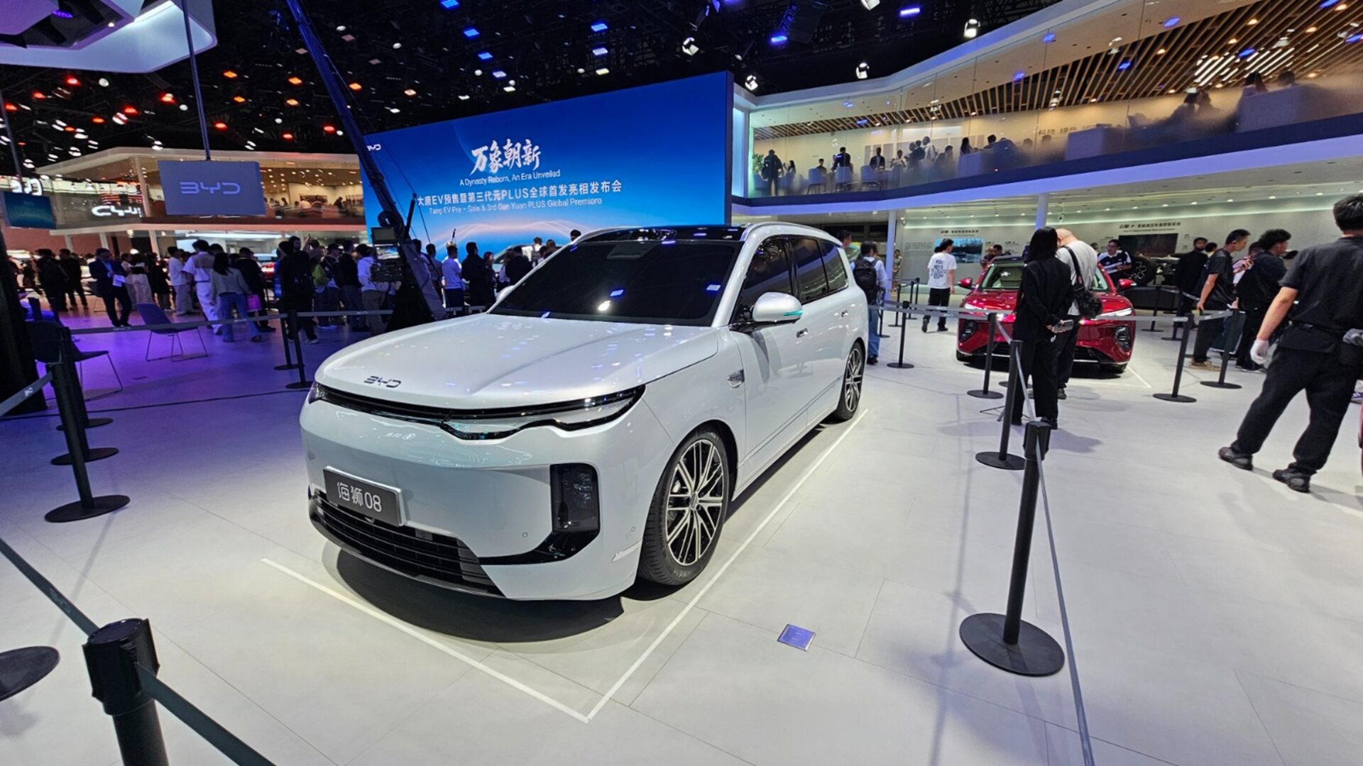 A white electric car displayed at an auto show, surrounded by attendees and other vehicles. The car features a modern design with a sleek front, prominent wheels, and is set against a backdrop with branding for BYD.