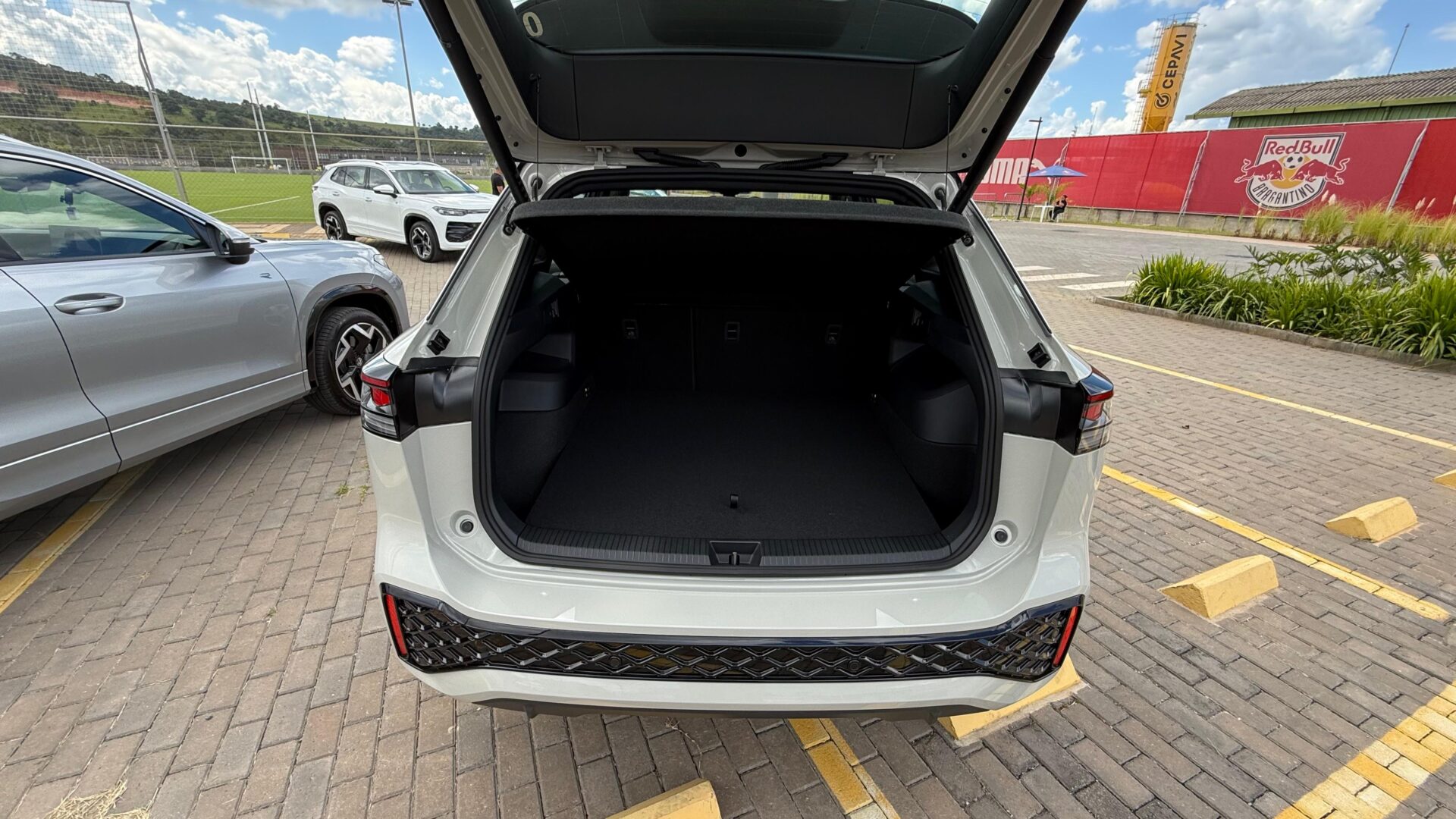 Imagem do porta-malas de um carro SUV aberto, mostrando o espaço vazio e o interior escuro. Ao fundo, há outros veículos estacionados e um campo de futebol.