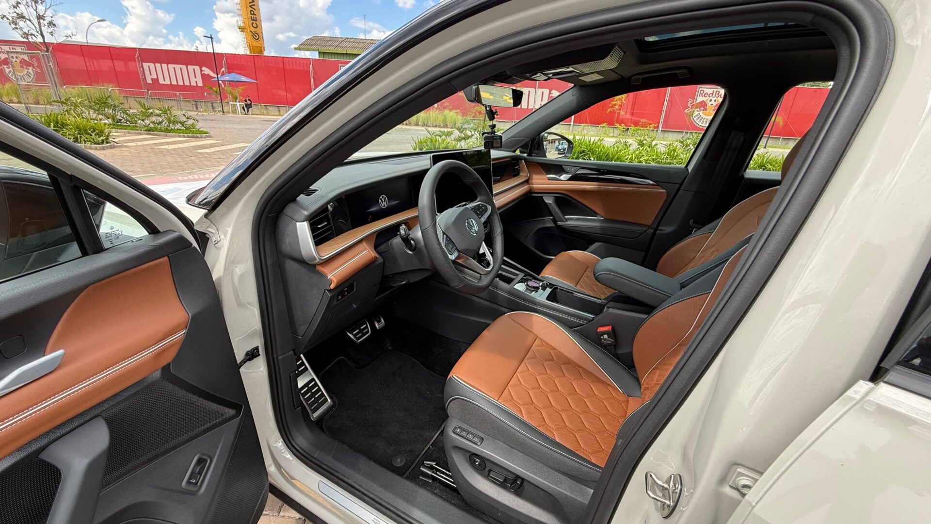 Interior de um carro moderno com bancos de couro marrom e detalhes em preto, volante com logo da Volkswagen, console central e painel com tela digital.