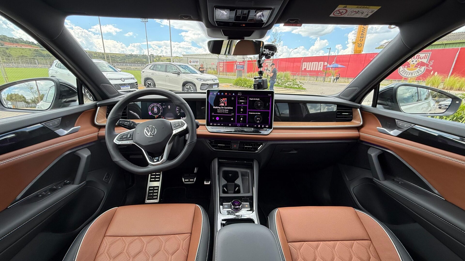 Interior de um carro Volkswagen com volante esportivo, painel digital, console central moderno e bancos em couro marrom.