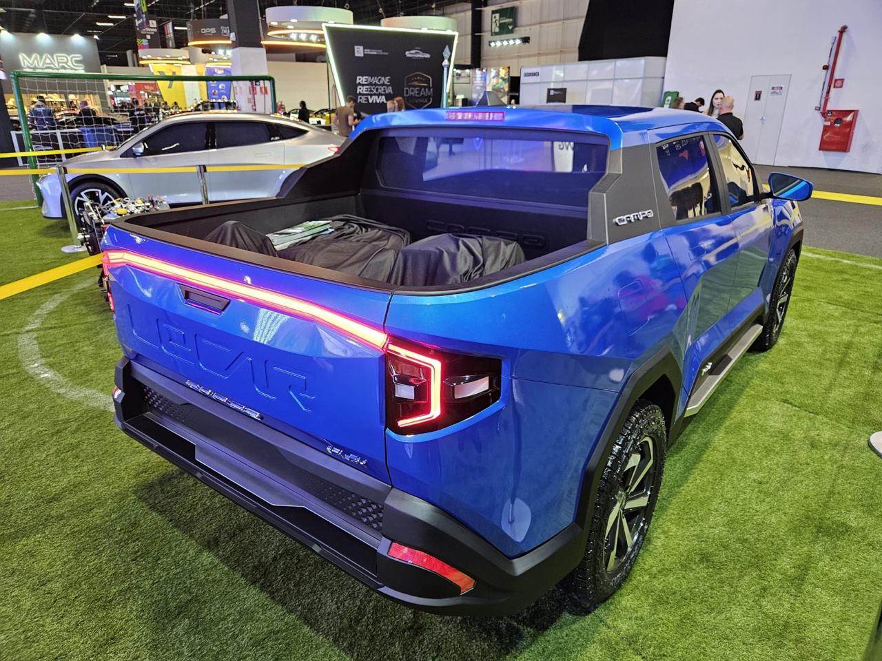 Imagem de um caminhão azul em uma feira de automóveis, com um design moderno e iluminação traseira em LED, estacionado sobre grama sintética.
