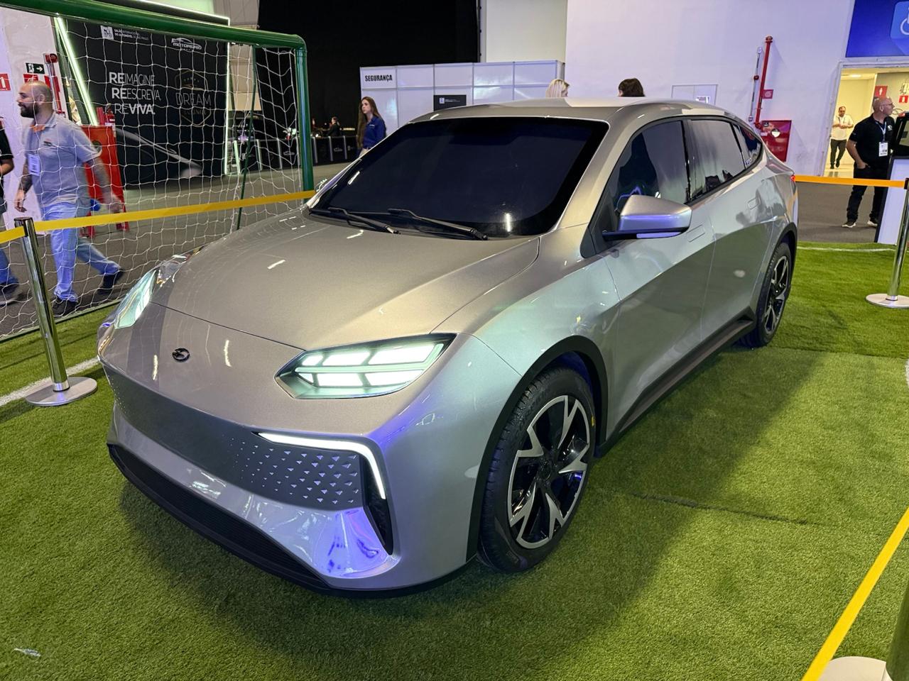 Carro elétrico prateado exibido em um evento com fundo de gramado e rede de futebol ao fundo.