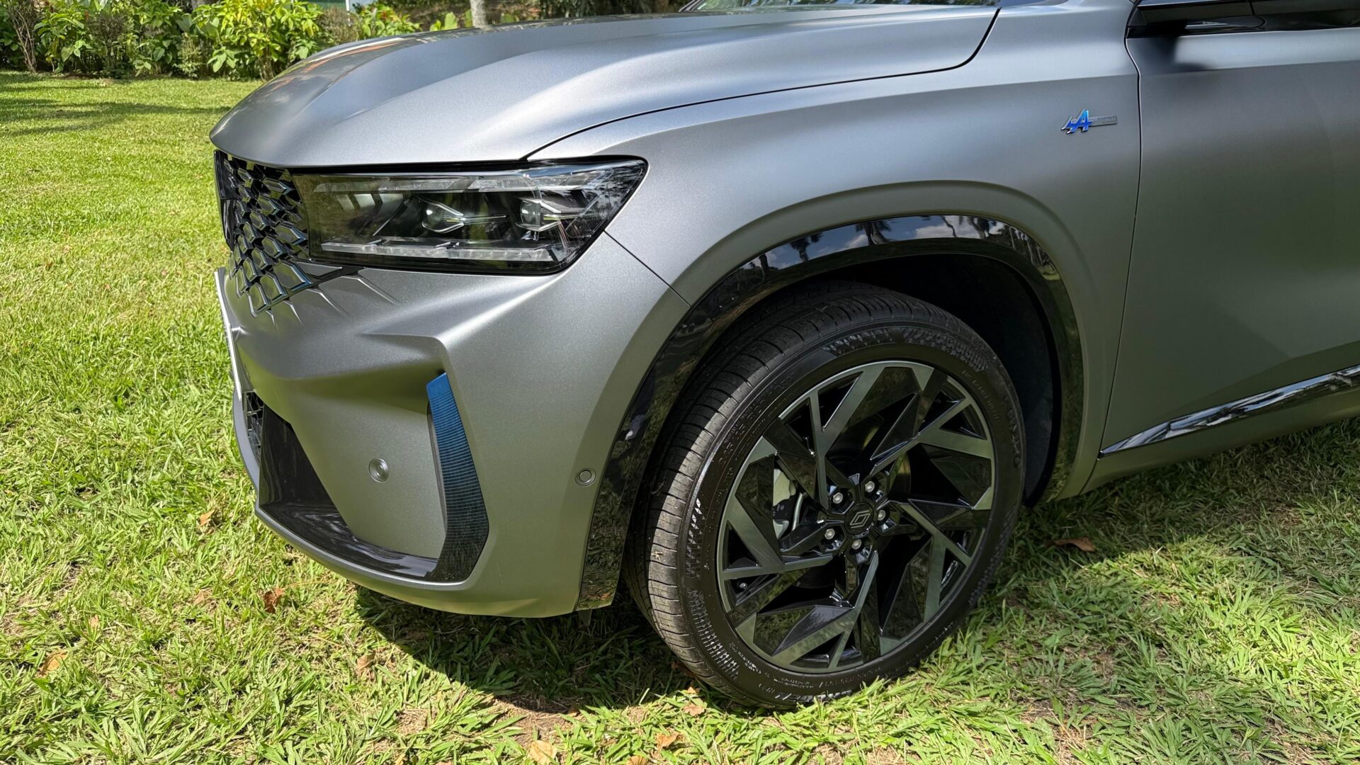 Detalhe do lado frontal de um carro cinza, mostrando a roda de liga leve preta e a parte do capô em um ambiente gramado.