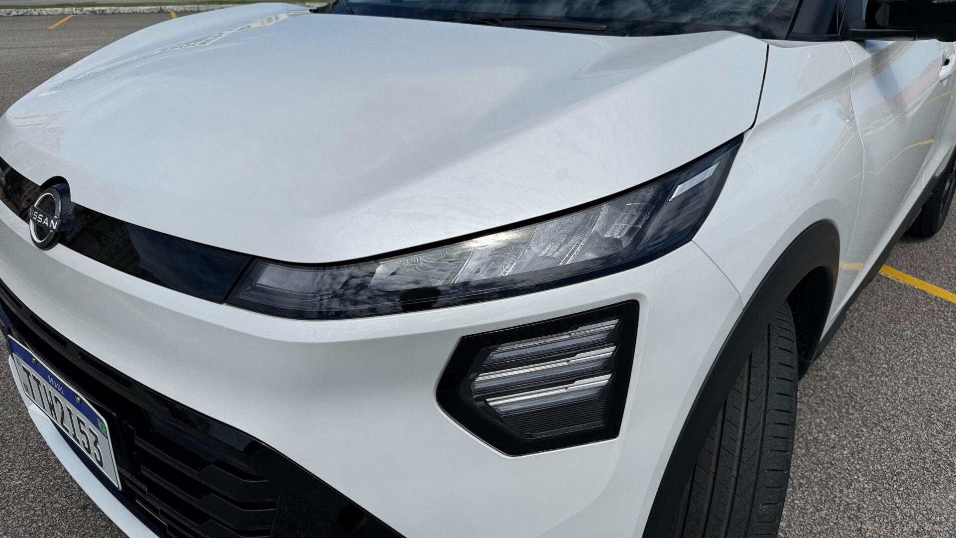 Close-up do capô e faróis de um carro Nissan branco em um estacionamento.