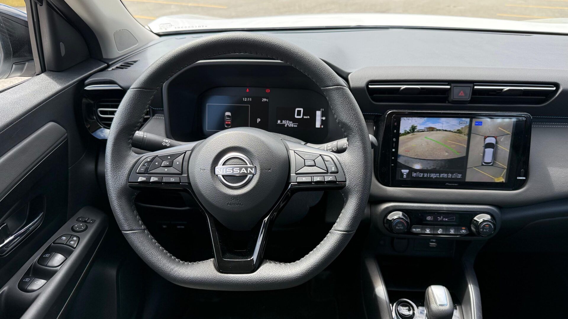 Interior de um carro Nissan, com foco no volante e painel de instrumentos. O volante possui botões de controle e o display central mostra uma câmera traseira.
